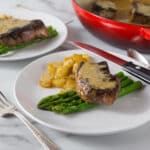 A photo of steak au poivre on a white place with asparagus and potatoes.
