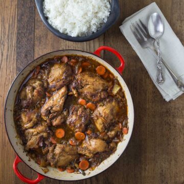 A red braising pan with Jamaican brown chicken stew and a black bowl with white rice.