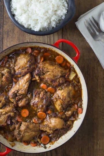 A red braising pan with Jamaican brown chicken stew and a black bowl with white rice.