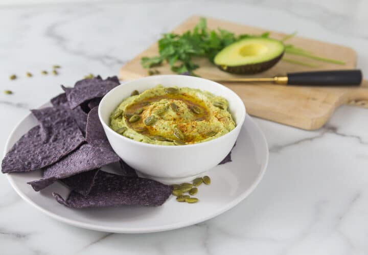 A photo of cannellini bean avocado hummus in a white bowl on a white plate with blue corn chips.