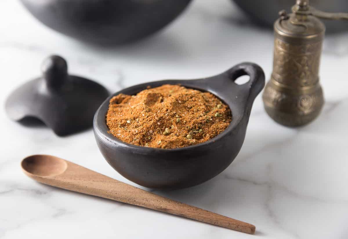 A photo of Turkish seasoning in a black ceramic bowl with a wooden spoon and spice grinder.