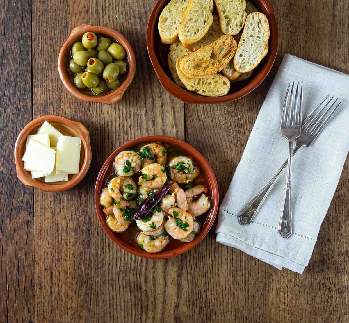 An overhead photo of gambas al ajillo in a dish and other dishes with bread, olives and cheese.
