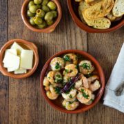 A photo of gambas al ajillo in a ceramic dish with other dishes with olives, cheese and bread.
