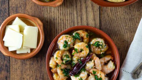 A photo of gambas al ajillo in a ceramic dish with other dishes with olives, cheese and bread.