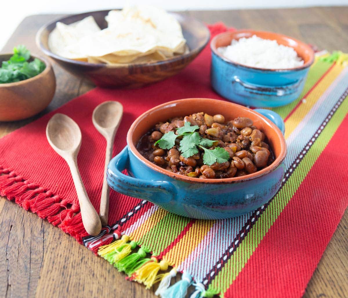 A photo of frijoles charros with cilantro, rice and tortillas in the background.