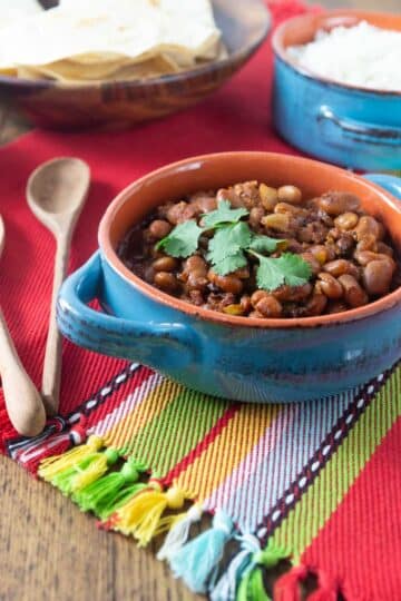 A photo of frijoles charros in a ceramic bowl with tortillas and rice in the background.