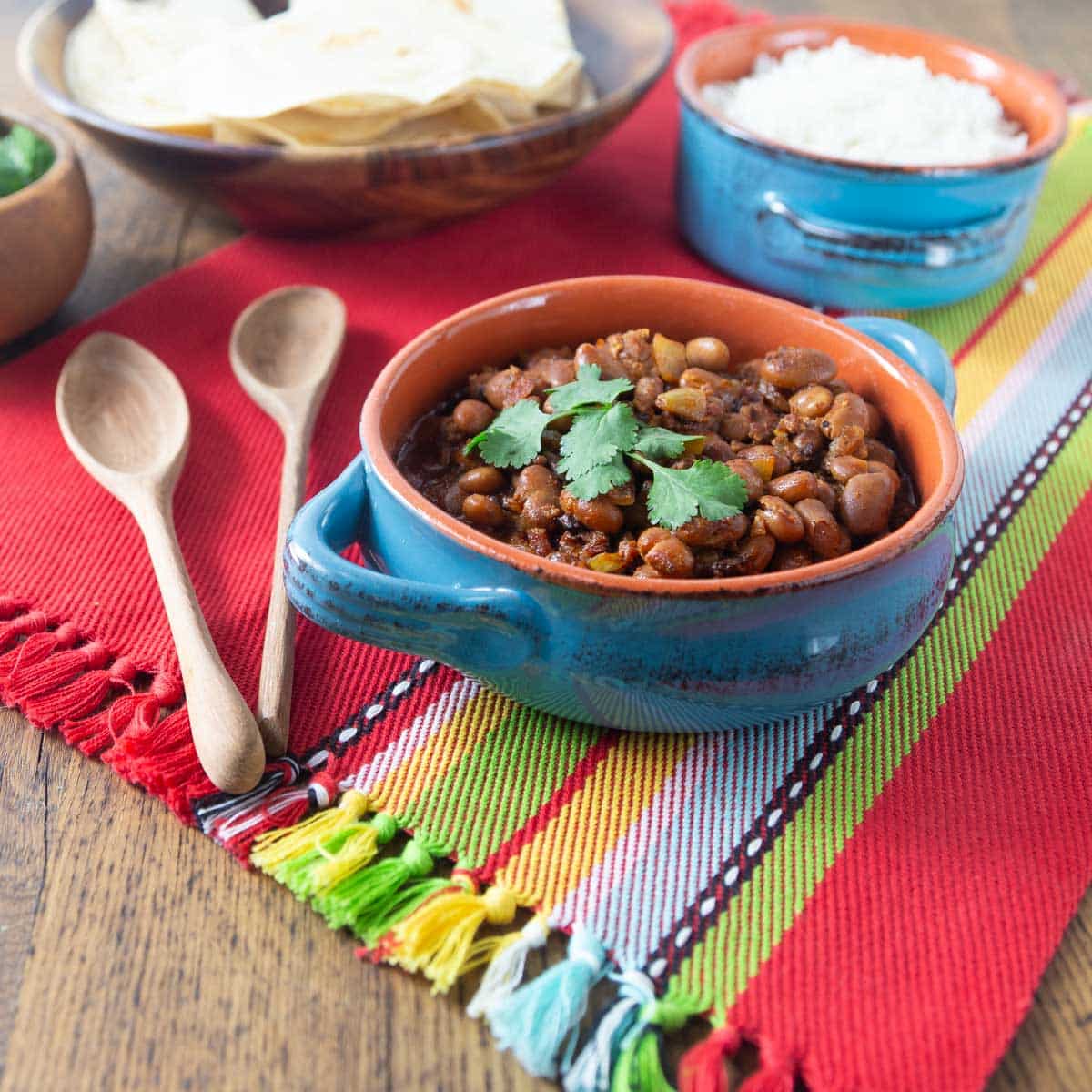 A photo of frijoles charros in a ceramic bowl with tortillas and rice in the background.