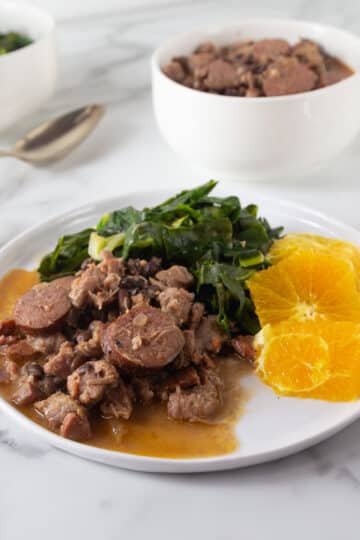 Brazilian feijoada on a white plate with collard greens and orange slices.