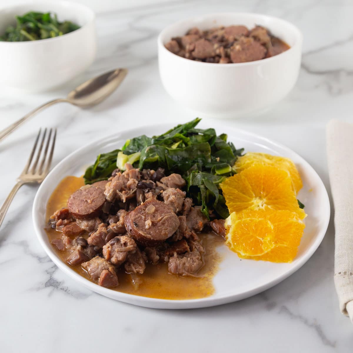 Brazilian feijoada on a white plate with collard greens and orange slices.