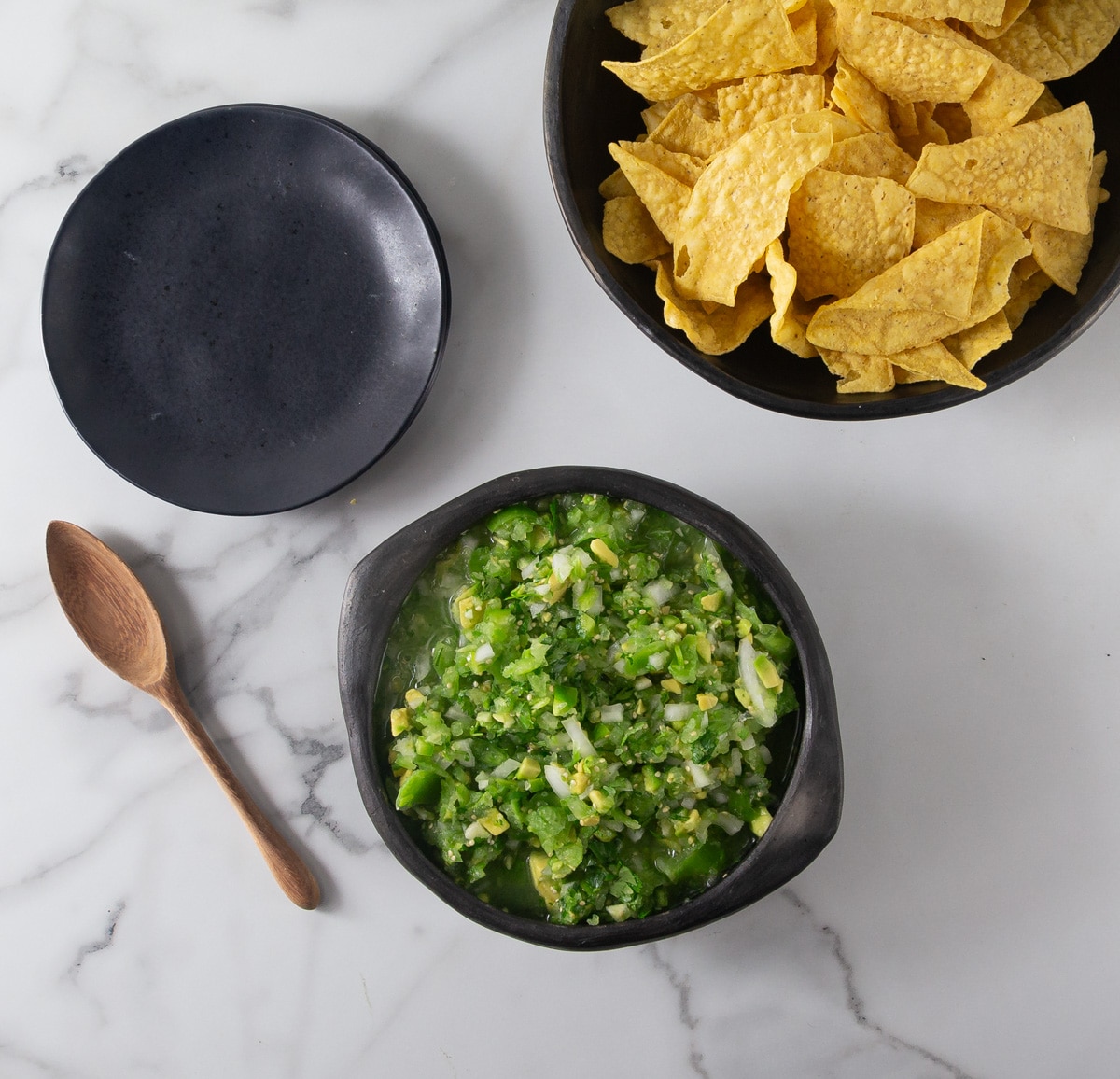 A photo of tomatillo avocado salsa with a spoon, black plates and chips.