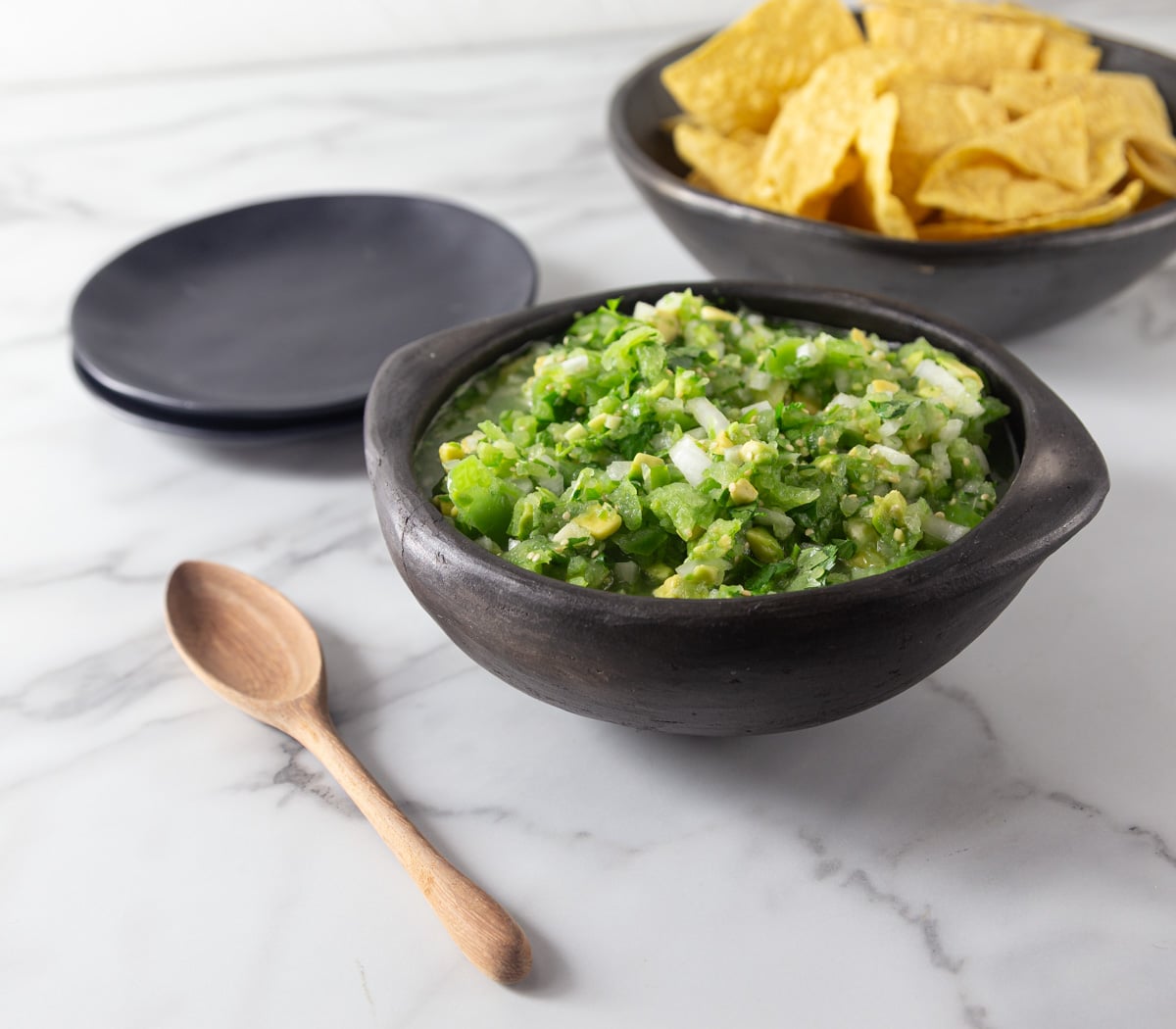 A photo of tomatillo avocado salsa with tortilla chips and plates in the background.