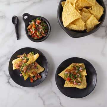 An overhead photo of pico de gallo in a black bowl with tortilla chips on plates.