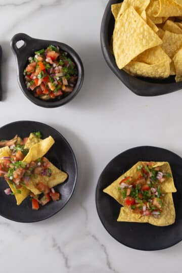 An overhead photo of pico de gallo in a black bowl with tortilla chips on plates.